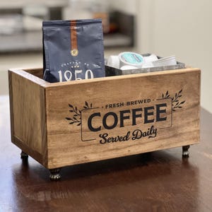 May include: A rectangular wooden storage box with four small metal feet. The front features black text that reads "FRESH BREWED COFFEE Served Daily" framed by leaf illustrations. Inside the box sits a dark blue coffee bag and a small metal container.