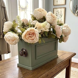 May include: A floral arrangement featuring large, cream-colored peonies and greenery in a sage green rectangular box. The box has a decorative handle and is placed on a wooden table. The flowers have a soft, natural appearance.