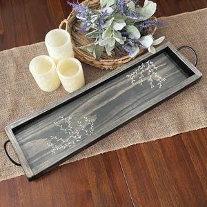 May include: A rectangular dark gray wooden tray with black metal handles on each end, featuring white painted sprig designs on the surface. Three off-white textured pillar candles and a woven basket containing lavender sprigs are nearby.