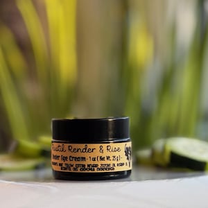 May include: A small black jar with a label that reads "Rustik Render & Rise Under Eye Cream - 1 oz (Net Wt. 25g)." The jar is sitting on a white surface with a blurred background of green plants.