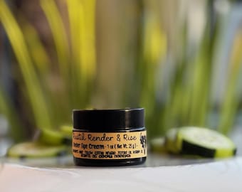 May include: A small black jar with a label that reads "Rustik Render & Rise Under Eye Cream - 1 oz (Net Wt. 25g)." The jar is sitting on a white surface with a blurred background of green plants.