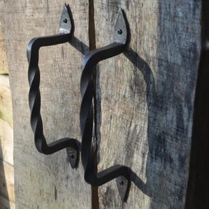 Può includere: Due maniglie per porta in ferro battuto nero montate su una porta in legno stagionato. Le maniglie presentano un design a torsione al centro e sono fissate con placche decorative a forma di foglia. La venatura del legno è visibile.