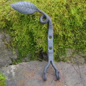May include: Hand-forged metal hook in the shape of a leaf. The hook is dark grey with a leaf design at the top, a curled stem, and two small hooks at the bottom. The background is green moss and a grey stone.