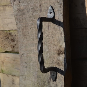 Pode incluir: Uma maçaneta de porta de ferro forjado à mão, preta, com um design central torcido. A maçaneta é montada numa porta de madeira castanha clara desgastada. As extremidades da maçaneta são fixadas com placas triangulares decorativas e parafusos.