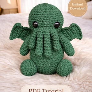 Może przedstawiać: Ręcznie robiona zielona szydełkowa pluszowa zabawka Cthulhu z czarnymi guzikowymi oczami, skrzydłami i mackami. Obraz zawiera tekst "Beginner-Friendly Pattern" i "PDF Tutorial". Widoczny jest również beżowy okrąg ze słowami "Instant Download".