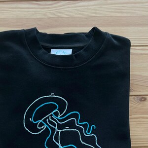 以下が含まれることがあります： 青と白の刺繍でクラゲのデザインが施された黒いスウェットシャツ。スウェットシャツには、「Ride the Wave」と書かれた小さなタグが前面についています。