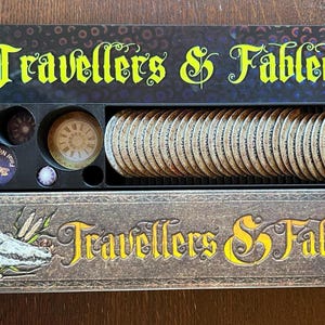 Puede incluir: Un organizador de juegos negro con el texto "Travellers & Fabled" en amarillo. El organizador contiene fichas y cartas de juego. Las fichas son redondas con varios diseños. Las cartas son beige con ilustraciones. El organizador está sobre una superficie de madera.
