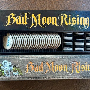 Puede incluir: Juego de mesa "Bad Moon Rising" con letras doradas. El juego incluye una bandeja negra con fichas redondas y montones de fichas blancas y negras. La caja inferior presenta un diseño de calavera y follaje.
