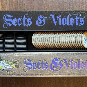 May include: A board game box set titled "Sects & Violets" in purple, decorative lettering. Inside the box are stacks of black tokens and a row of circular game pieces. The bottom box features a skull and floral design.