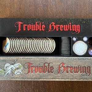 May include: A board game box set titled "Trouble Brewing" in red script. The box contains game pieces, including circular tokens and a stack of black cylindrical pieces. The box has a rustic, vintage aesthetic.