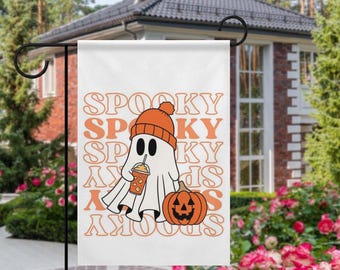 May include: White Halloween garden flag featuring a cartoon ghost wearing an orange beanie and holding a pumpkin spice drink. A jack-o'-lantern and the word "Spooky" in orange text are also on the flag. The flag is hanging in a garden.