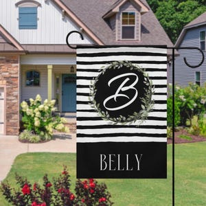 Puede incluir: Una bandera de jardín a rayas horizontales en blanco y negro. Presenta un diseño circular con la letra blanca "B" rodeada por una corona de hojas verdes. La palabra "BELLY" se muestra en letras blancas en la parte inferior.