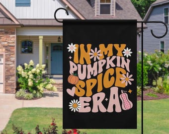 May include: Black garden flag with the text "In My Pumpkin Spice Era" in orange, pink, and white. The flag features a pumpkin and floral designs. The flag is hanging in front of a house.
