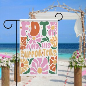 May include: A garden flag displaying the text "FDT AND HIS SUPPORTERS" in orange, green, and yellow, framed by pink, orange, and yellow floral designs. The flag is suspended from a black metal stand, set against a beach and ocean backdrop.
