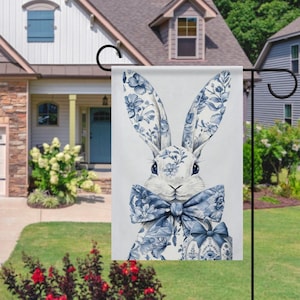 May include: A garden flag with a watercolour illustration of a white rabbit adorned with blue floral patterns on its ears, bow tie, and an Easter egg. The flag is displayed outdoors.