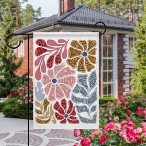 Puede incluir: Una bandera de jardín blanca con un diseño floral. Las flores y las hojas están llenas de las palabras "FUCK TRUMP" en varios colores, incluyendo rojo, marrón y azul. La bandera está colgada en un soporte de metal negro en un jardín.