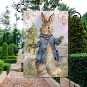May include: A garden flag with a watercolour illustration of a rabbit in a blue coat, holding a leafy branch. The background shows a house, butterflies, and script. The flag is displayed on a black metal stand.