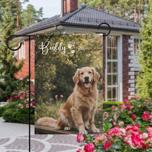 May include: A garden flag with a Golden Retriever dog on a bench. The flag has the name "Buddy" and paw prints. The background shows a blurred green landscape and a house with a red brick exterior. The flag is displayed in a garden.