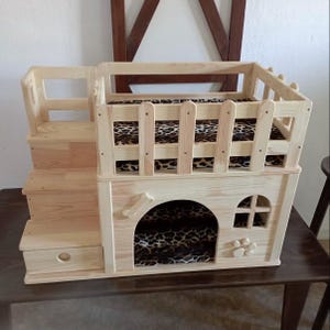 Puede incluir: Una casa de madera para mascotas con escalera y plataforma elevada. La casa presenta una cama con estampado de leopardo y un tirador en forma de hueso. El diseño incluye una pequeña ventana y detalles en forma de pata.