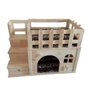 Puede incluir: Una casa de mascotas de madera con un diseño de varios niveles. Cuenta con una escalera, un pequeño balcón con una valla y un interior acogedor con un cojín con estampado de leopardo. La casa incluye un recorte en forma de hueso de perro y un diseño en forma de flor.