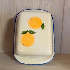Puede incluir: Una mantequera de cerámica con tapa, con base blanca y borde azul. La tapa está decorada con dos ilustraciones de cítricos naranjas con hojas verdes. El plato está diseñado para uso en la cocina.