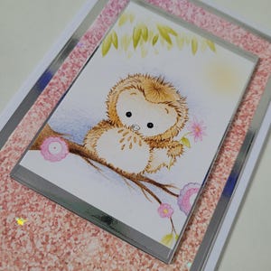 May include: A greeting card with a charming cartoon owl illustration. The owl, with fluffy brown fur, sits on a branch and holds a pink flower. The card has a pink glitter border and a silver frame, with a soft, light background.