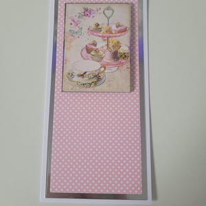 May include: A decorative card with a vintage image of a tiered cake stand with cupcakes and a teacup. The card has a pink polka dot background and a silver border, creating a sense of elegance and charm.