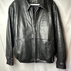 May include: A black leather jacket with a full-length front zipper, a pointed collar, and two side pockets. The jacket features long sleeves with buttoned cuffs and a quilted interior lining. The brand label reads Colebrook.