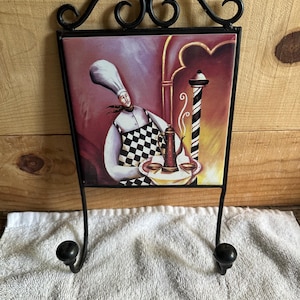 May include: Black metal wall decor featuring a ceramic tile with a chef illustration. The chef wears a tall white hat and checkered apron. The background has a barber pole and a coffee pot. Two black hooks are below the tile.