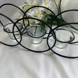 May include: Black metal candle holder with three clear glass votive cups. The holder has a looping design and is decorated with white flowers and green leaves.