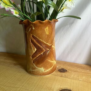 May include: A ceramic vase with a brown glaze and a decorative chevron design. The vase has a ruffled rim and is filled with artificial white and yellow flowers with green stems and leaves. The vase is sitting on a wooden surface.