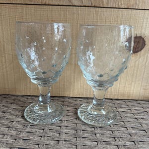 Könnte beinhalten: Zwei klare Weingläser mit strukturierter Oberfläche. Die Gläser haben einen Stiel und einen runden Fuß. Der Kelch und der Fuß der Gläser haben eine blasenartige Textur. Die Gläser werden auf einer gewebten Oberfläche mit einem Holzgrund gezeigt.