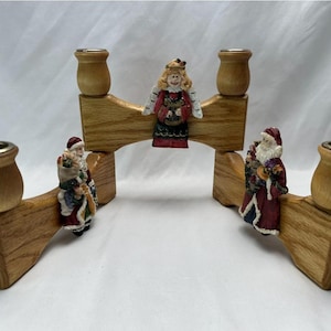 May include: Wooden candle holders with decorative figurines of Santa Claus and an angel. The candle holders are made of light brown wood and have gold-colored metal candle cups. The figurines are colorful and add a festive touch.