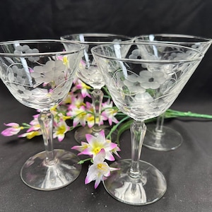 Könnte beinhalten: Ein Set aus vier klaren Cocktail-Coupes aus Glas mit einem zarten, geätzten Blumenmuster auf der Schale. Jedes Glas hat einen schlanken Stiel und einen runden Fuß und besticht durch ein transparentes Design mit feinen, gravierten Blumendetails.
