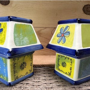 Op de afbeelding: Twee decoratieve keramische dozen met een lampachtig ontwerp. Elke doos heeft een blauwe basis en bovenkant, met witte zijkanten met bloemen- en geometrische patronen in geel, groen en blauw. De dozen zijn met de hand beschilderd in een grillige stijl.