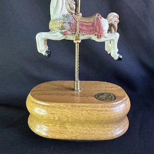 Pode incluir: Estatueta de cavalo de carrossel com corpo branco, crina e cauda castanhas e freio vermelho. O cavalo tem uma sela rosa e é montado em uma base de madeira com um poste dourado.