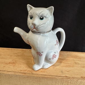 Puede incluir: Una tetera de cerámica blanca con forma de gato sentado. El gato tiene rasgos faciales pintados que incluyen ojos azules, bigotes negros y una boca roja. El cuerpo está decorado con pequeños patrones florales rojos repetidos sobre la superficie.