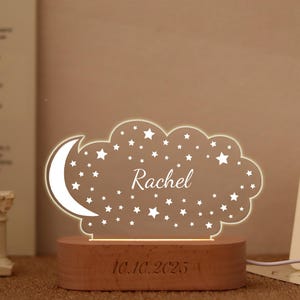 May include: A cloud-shaped acrylic night light featuring a crescent moon and star pattern with the name Rachel engraved in the center. The light is mounted on a rectangular wooden base with the date 10.10.2025 engraved on the front surface.