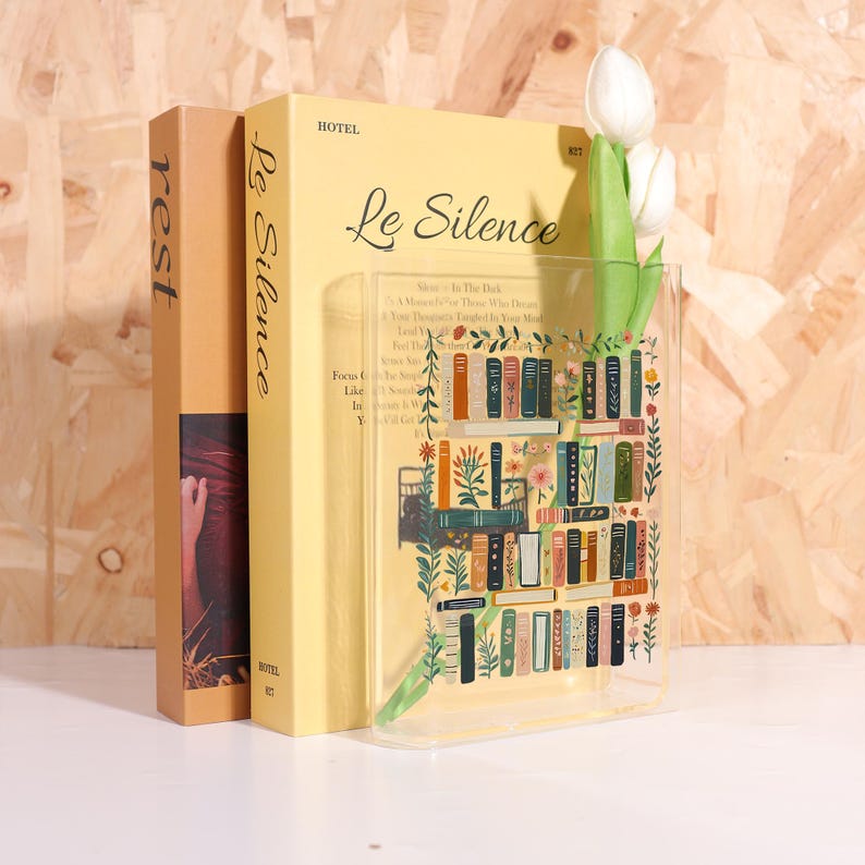Pode incluir: Um vaso de acr&iacute;lico transparente com um design de estante, segurando duas tulipas brancas. O vaso est&aacute; posicionado em frente a dois livros, um intitulado "Le Silence" e o outro "rest". Os livros s&atilde;o em tons de amarelo e castanho.