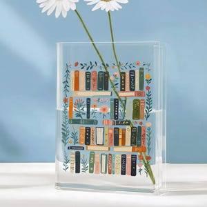 Pode incluir: Vaso retangular de acr&iacute;lico transparente com uma ilustra&ccedil;&atilde;o colorida de uma estante cheia de livros e detalhes florais. O vaso cont&eacute;m duas flores de margarida brancas. O fundo &eacute; azul claro. Altura aproximada: 20 cm.