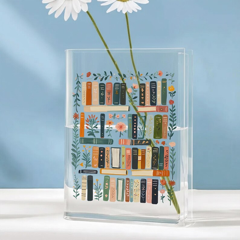 Pode incluir: Vaso retangular de acr&iacute;lico transparente com uma ilustra&ccedil;&atilde;o colorida de uma estante cheia de livros e detalhes florais. O vaso cont&eacute;m duas flores de margarida brancas. O fundo &eacute; azul claro. Altura aproximada: 20 cm.