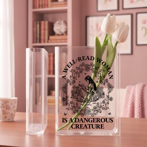 May include: A clear rectangular acrylic vase featuring a black graphic print of a person reading surrounded by flowers and the text A WELL-READ WOMAN IS A DANGEROUS CREATURE. Two white tulip stems are placed inside the transparent container.