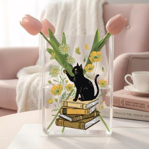 May include: Clear acrylic vase with a black cat illustration sitting on a stack of books. The vase is decorated with yellow flowers and butterflies, and holds pink tulips. A charming and decorative floral vase.