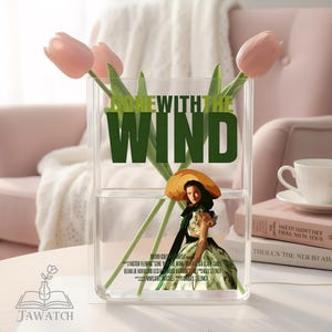 May include: A clear rectangular acrylic vase featuring the green text GONE WITH THE WIND and a graphic illustration of a person in a wide-brimmed hat and dress. Three light pink tulips with green stems are arranged inside the transparent container.