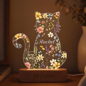 May include: A cat-shaped night light with a floral design and the name "Rachel" etched into the acrylic. The light sits on a wooden base with the date "07.15.2024" engraved. The light emits a warm glow.