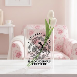 May include: A clear rectangular glass vase featuring a black graphic print of a person reading among books, surrounded by a floral wreath. The text reads A WELL-READ WOMAN IS A DANGEROUS CREATURE. Two white tulip stems are placed inside the vase.