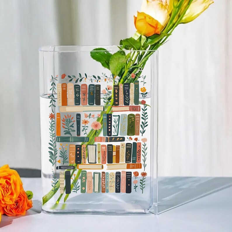 Pode incluir: Um vaso de vidro transparente e retangular com um design floral e de estante de livros. O vaso cont&eacute;m &aacute;gua e uma rosa amarela com folhas verdes. O design da estante apresenta lombadas de livros em tons de castanho, verde e laranja.
