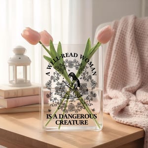 May include: A clear rectangular glass vase featuring a black graphic print of a person reading among books and flowers, with the text A WELL-READ WOMAN IS A DANGEROUS CREATURE. The vase contains three light pink tulips with green stems and leaves.