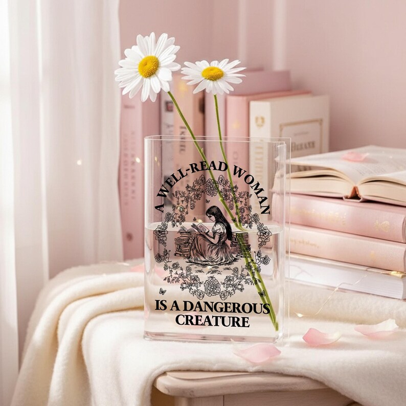 May include: A clear rectangular acrylic vase featuring a black graphic illustration of a person reading among flowers. The text A WELL-READ WOMAN IS A DANGEROUS CREATURE is printed in black. Two white daisies with yellow centers are placed inside the vase.