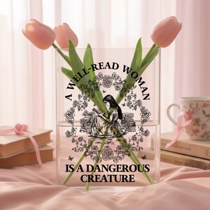 May include: A clear rectangular acrylic vase featuring a black graphic print of a person reading among stacks of books and floral wreaths. The text reads A WELL-READ WOMAN IS A DANGEROUS CREATURE. Three pink tulips are placed inside the vase.